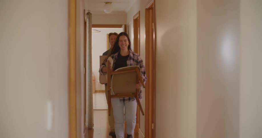 A man and woman smile as they walk down a hall carrying a chair and a box