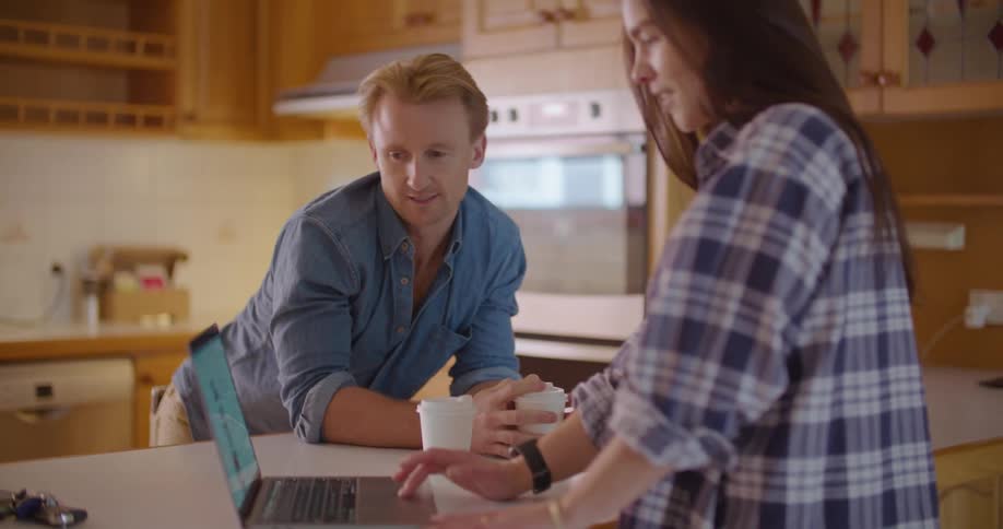 A man and woman stand in a kitchen with cups of coffee and talk and look at a laptop