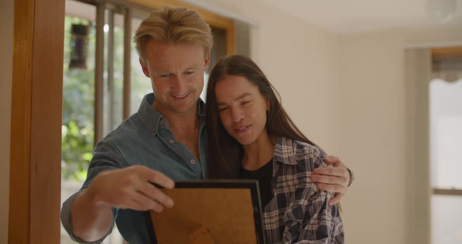Blonde man with a blonde shirt holds woman and takes a photo frame out of a box in the living room