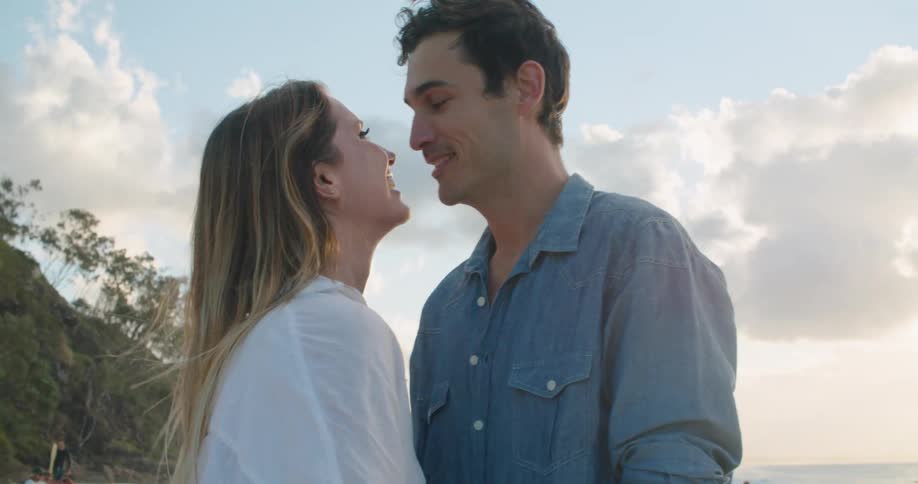 A man and a woman standing on a beach holding hands and kissing with the ocean sky and clouds in the background