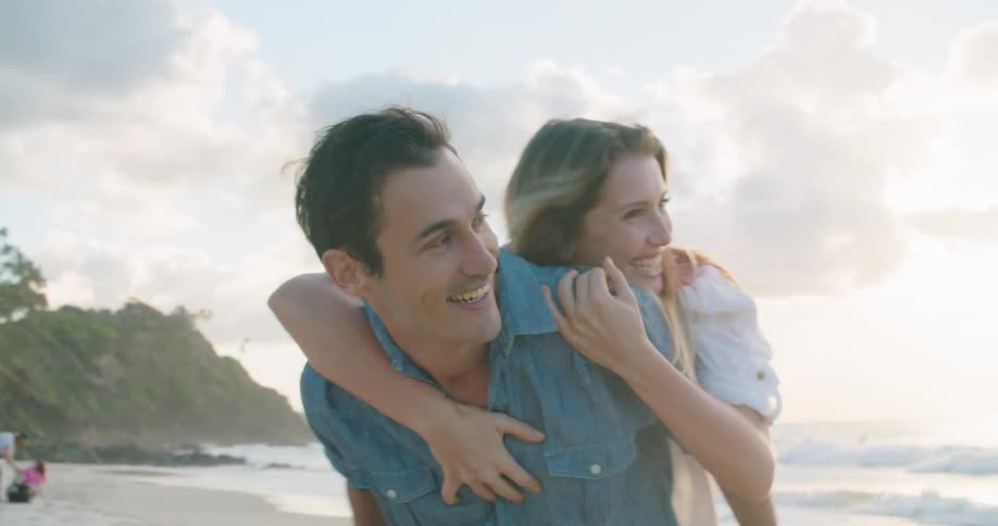 Man in a blue shirt carries woman with a white shirt on his back on the beach
