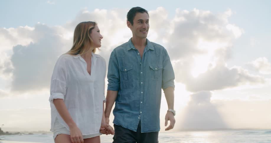 Young couple walk holding hands on the sand beach near the ocean at sunset