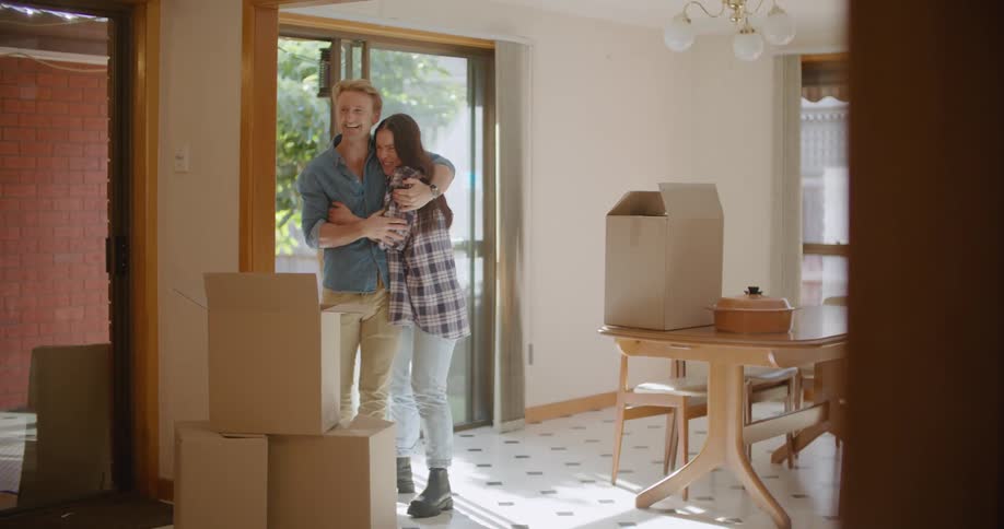 A woman carries in a box then hugs her husband as they look around and enjoy moving in