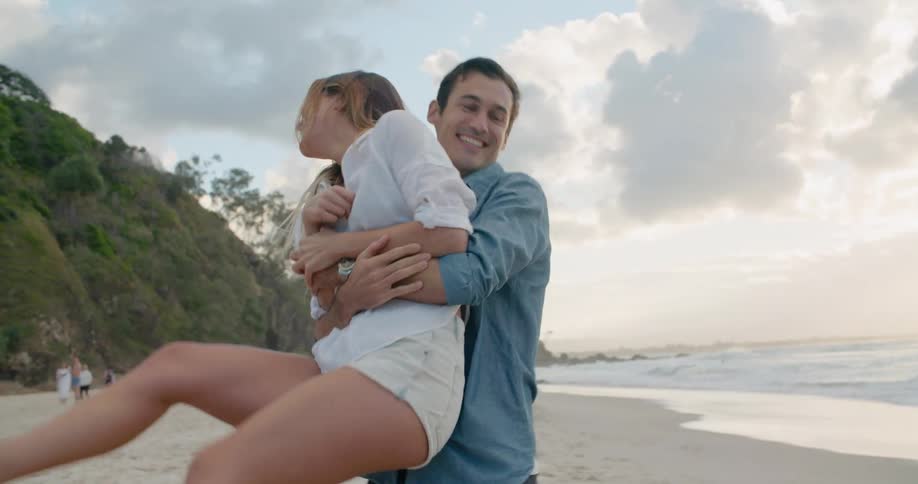 A man lifts a woman as they are smiling on a beach next to the ocean