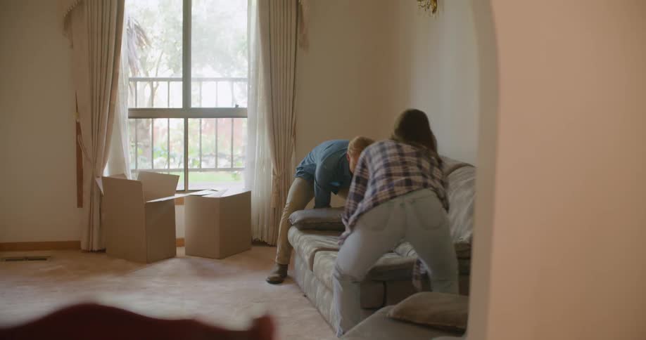 A man and woman carry a sofa across the room and set it down before they both sit down atop it