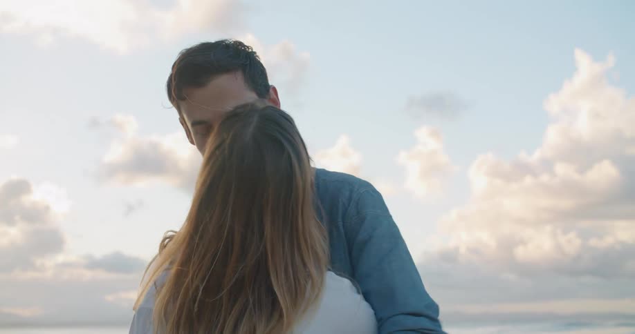 A man and a woman holding hands and hugging in a beach with the sky and clouds in the background