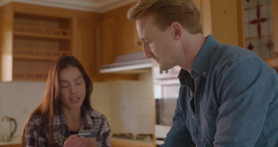 Man types on laptop standing in kitchen as woman holds credit card and talks