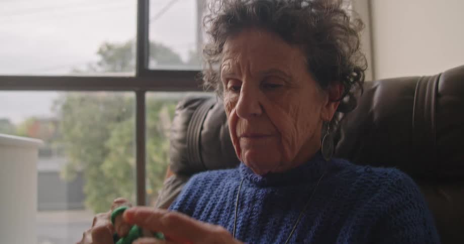 A woman knits with green yarn while sitting in a recliner next to her window