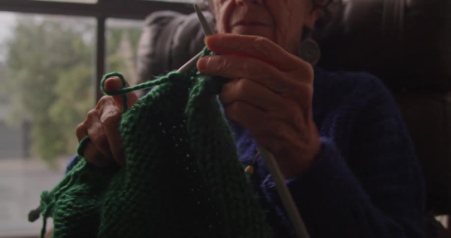 A woman sits next to a window in a recliner and knits using green yarn