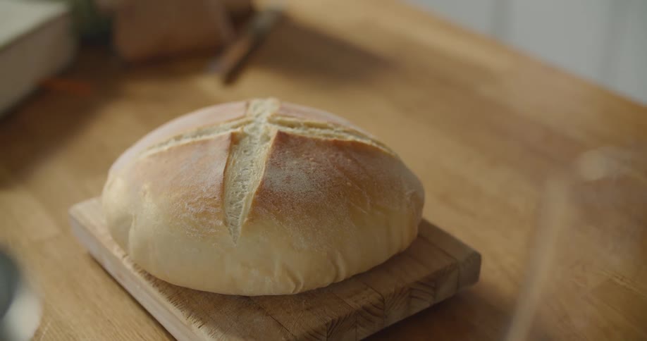 Person unwraps oval loaf from paper and places it on wooden chopping board on wooden table