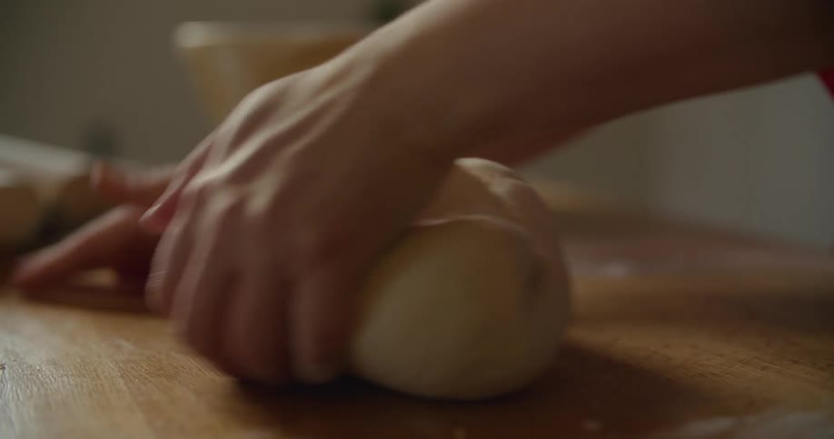 Person kneads dough on wooden surface near yellow bowl and open cookbook