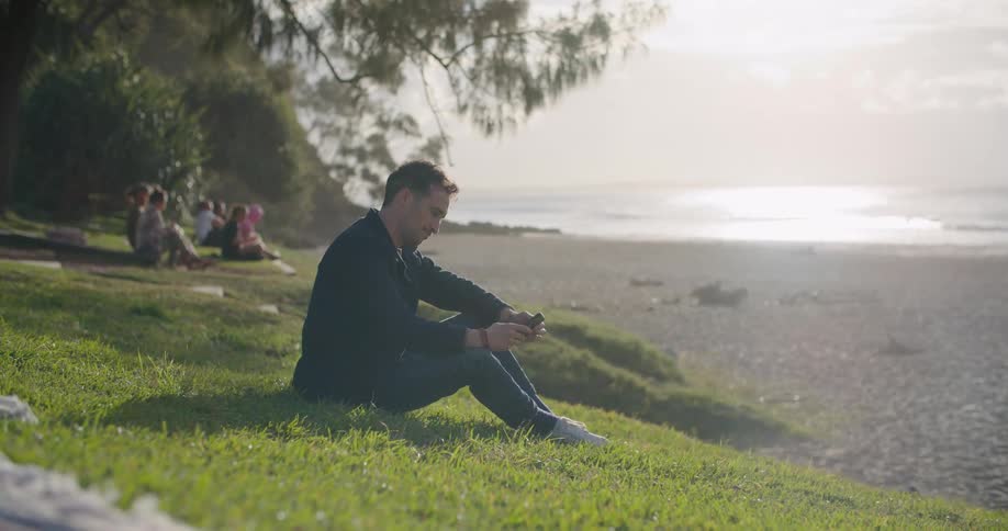 A man sitting near a beach using a smartphone