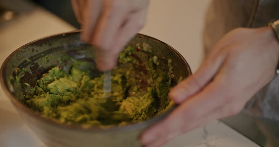 A woman mashes avocado in a bowl while a man browns ground beef in a pan