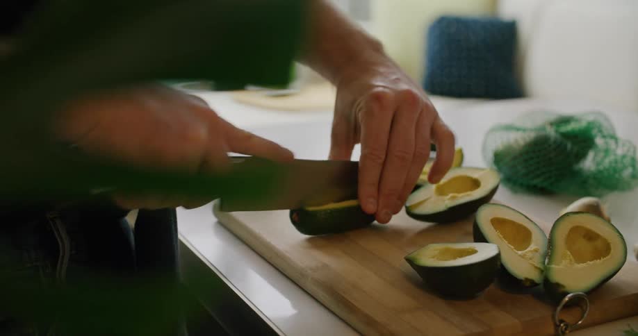 A male uses a large knife to cut multiple avocados into smaller slices