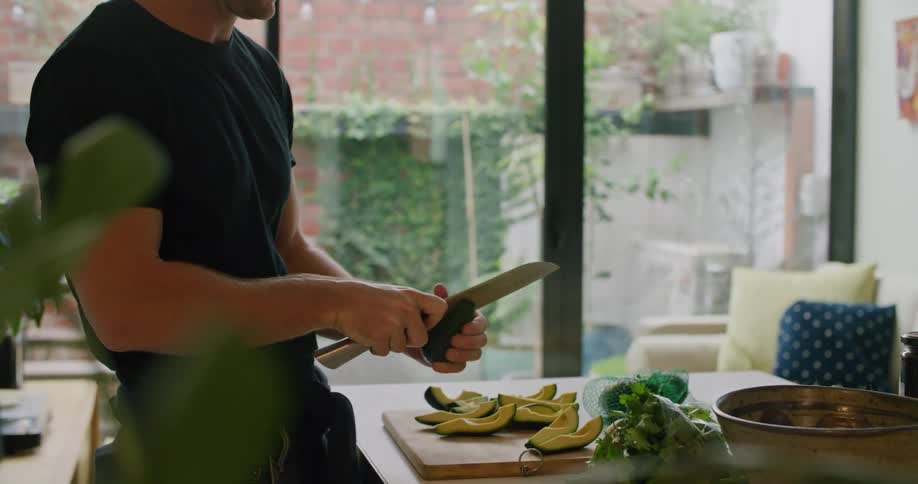 A man stands in his kitchen while he slices avocado with a large knife