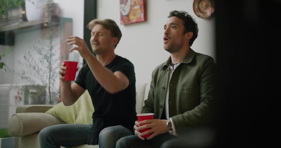 Two men sit on a couch with red solo cups and laugh and point as they look at something