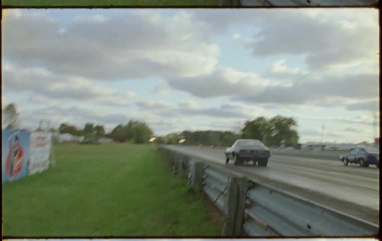 Two vehicles drag racing on a racetrack with trees the sky and clouds in the background