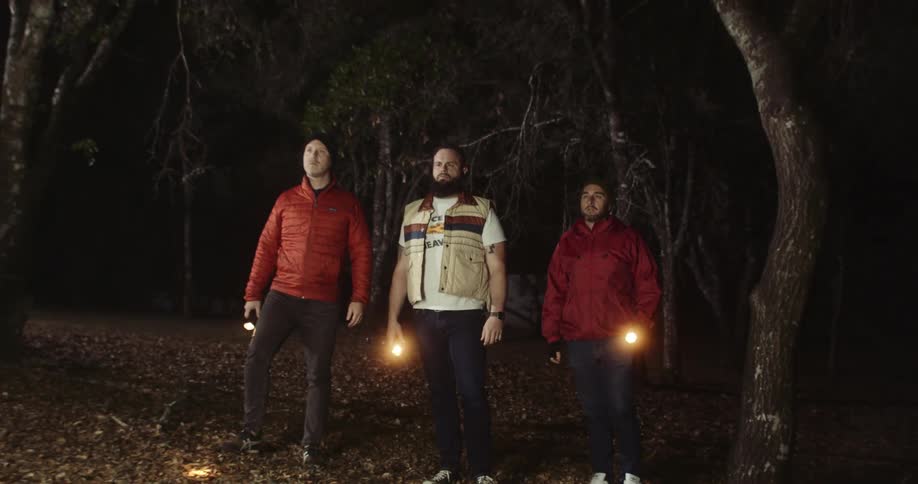 Three men carrying flashlights walk through woods approaching glowing rectangular object