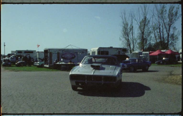 A line of sports cars drive through a campground filled with recreational vehicles