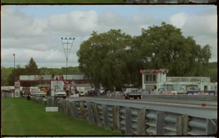 Two vehicles drag racing on a track with trees the sky and clouds in the background