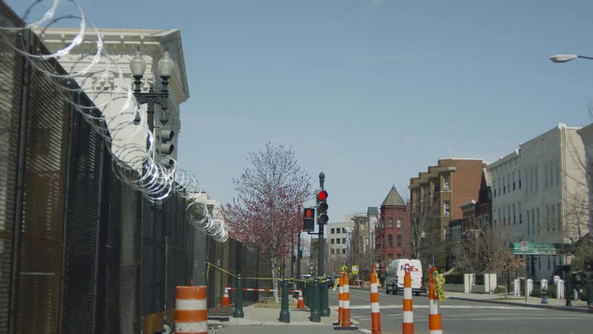 Pedestrians walks along sidewalk near intersection with red light across street from fence topped with razor wire
