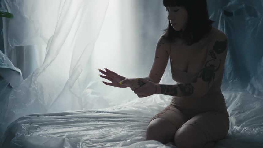 Woman kneeling on plastic draped platform surround by plastic defusing light from window examines hand with long nails