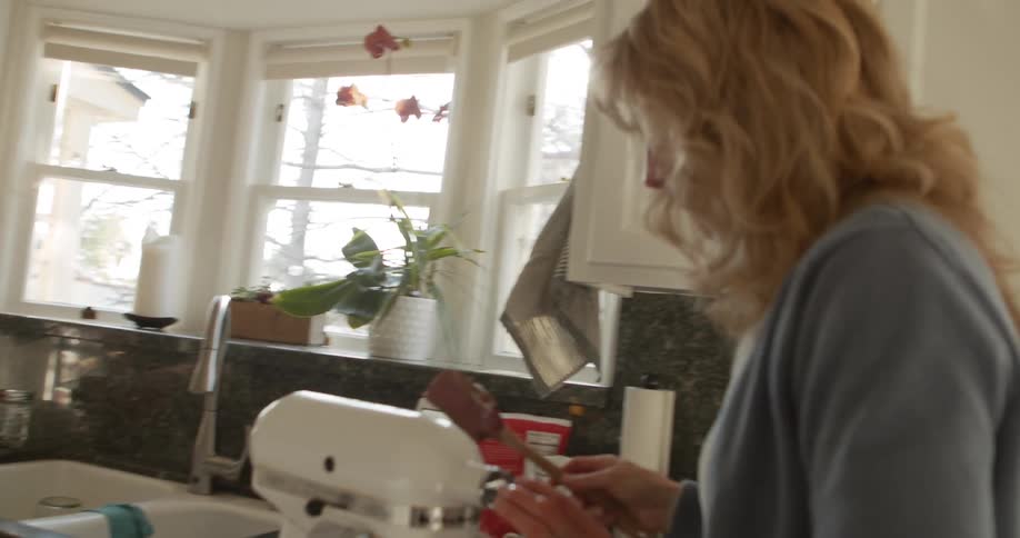 A woman carries a spatula with chocolate frosting on it while she checks on her electric mixer
