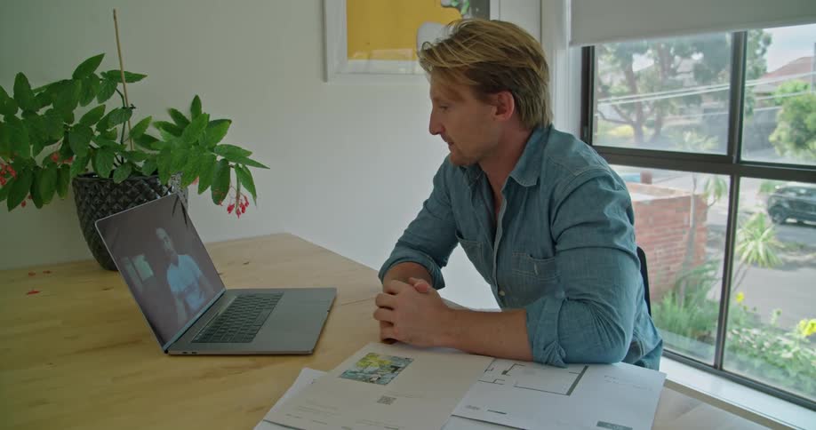 A man raises his hand to greet a colleague on the laptop screen while sitting at his desk in an office