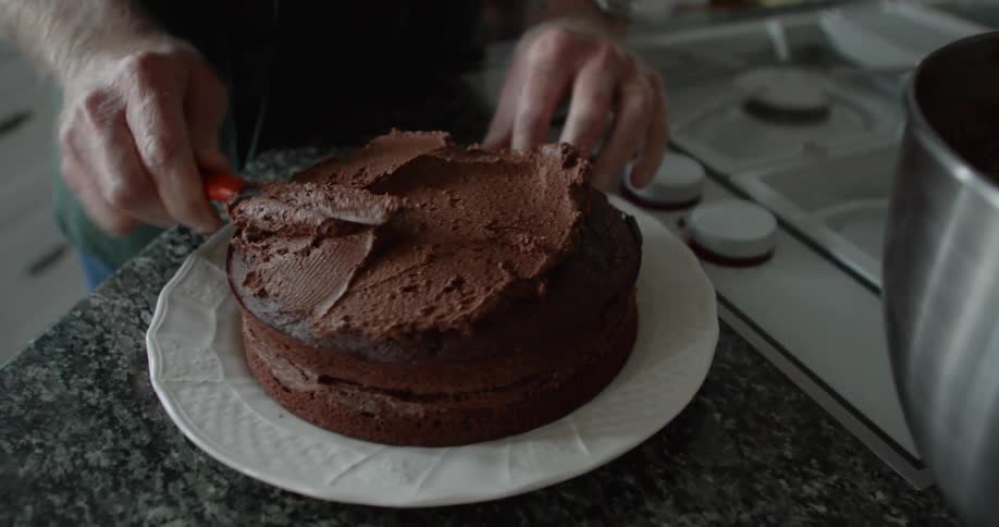 A person in a kitchen applying frosting to a cake with cabinets in the background