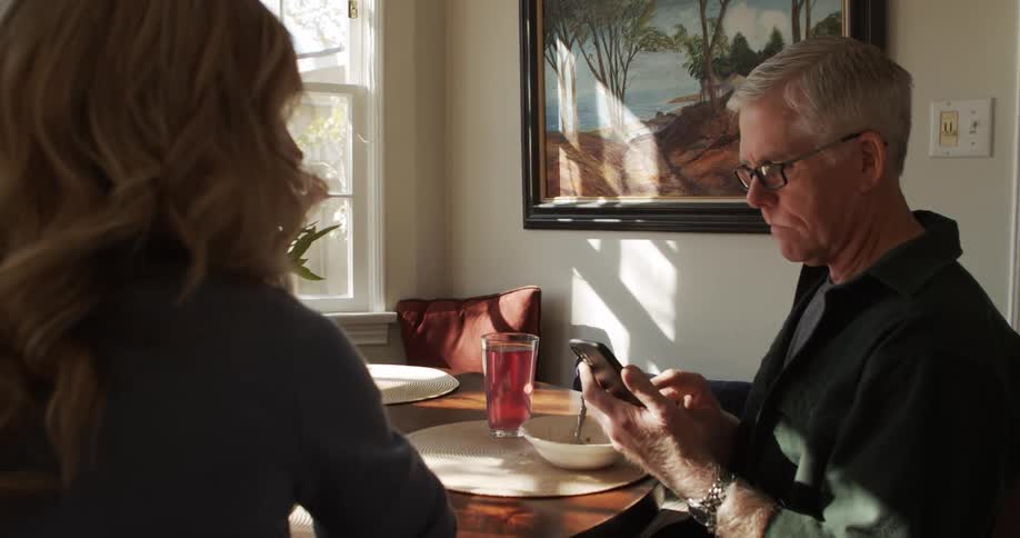 Man with glasses and a shirt and blonde woman sit at table uses smartphone in the kitchen