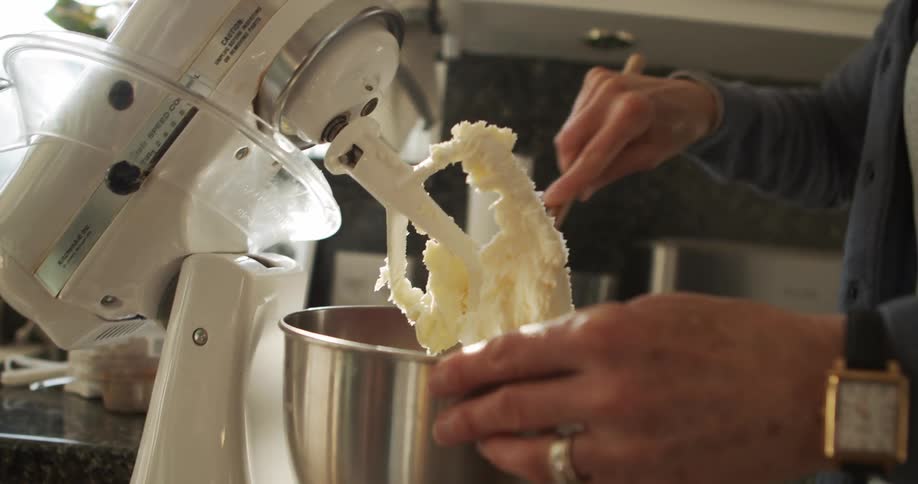 A woman uses a spatula to scrape dough from the agitator of a mixer in a home kitchen during the day