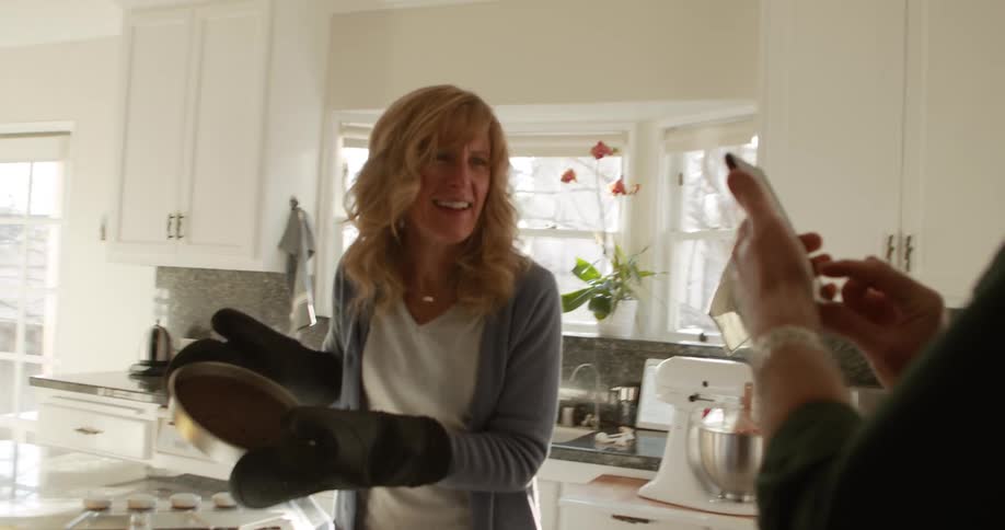 A man takes a photo of a woman with his cell phone while she holds up a cake she baked