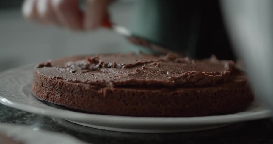 A person evenly spreads chocolate frosting over the top of a layer of cake