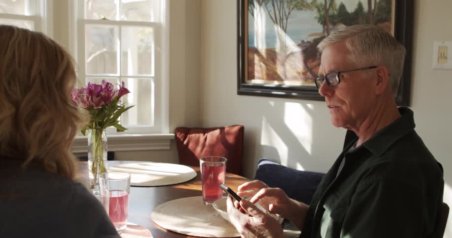 A man scrolls through his smartphone while having lunch at home with his wife