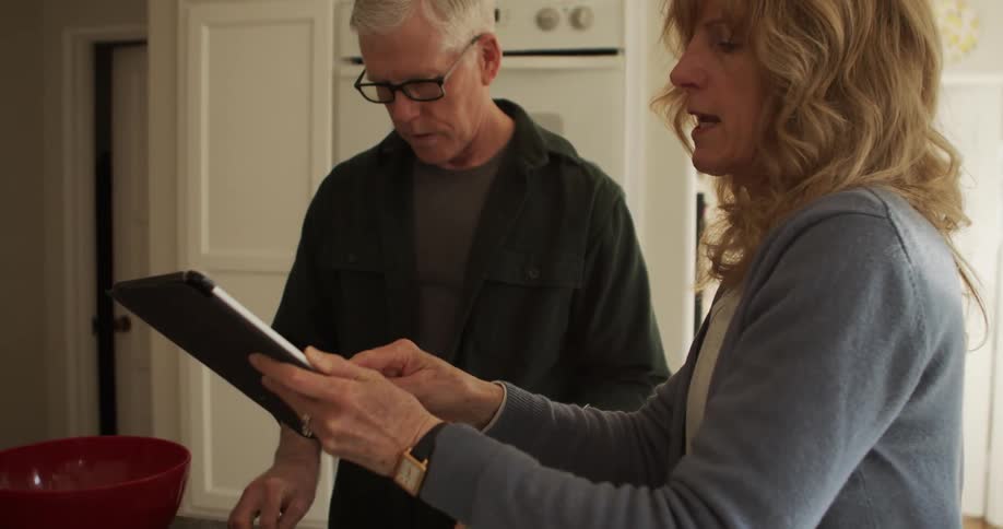 A man and woman stand in a kitchen while the woman reviews a recipe on her tablet computer