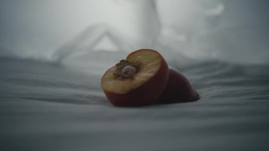 A single peach that is cut in half lay on a sheet as thin fabric dangles behind it