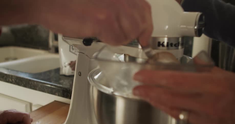 A man adds ingredient to a standing mixer with a measuring cup as his wife stands with spatula in her hand