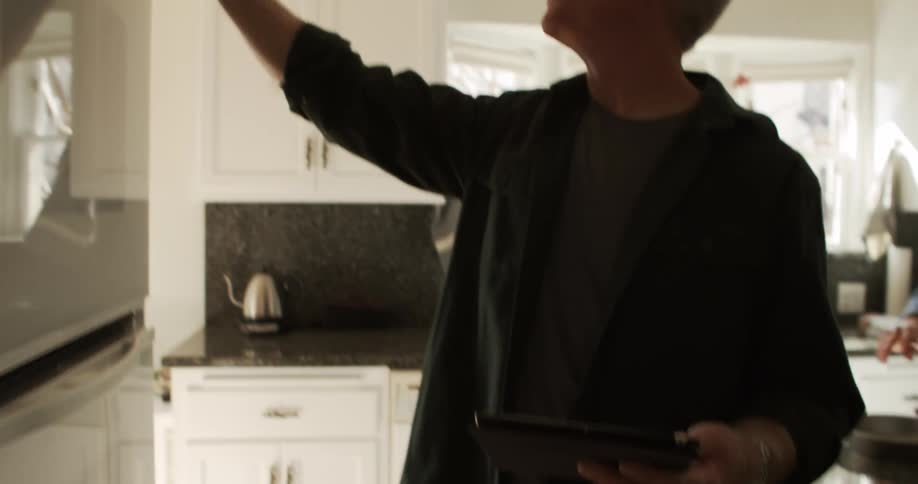 A man holding a tablet turns a knob on an oven while standing in a home kitchen during the day
