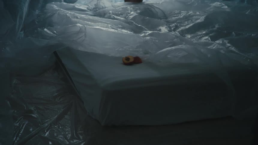 A person lies motionlessly atop a large mattress while covered in a sheet of plastic