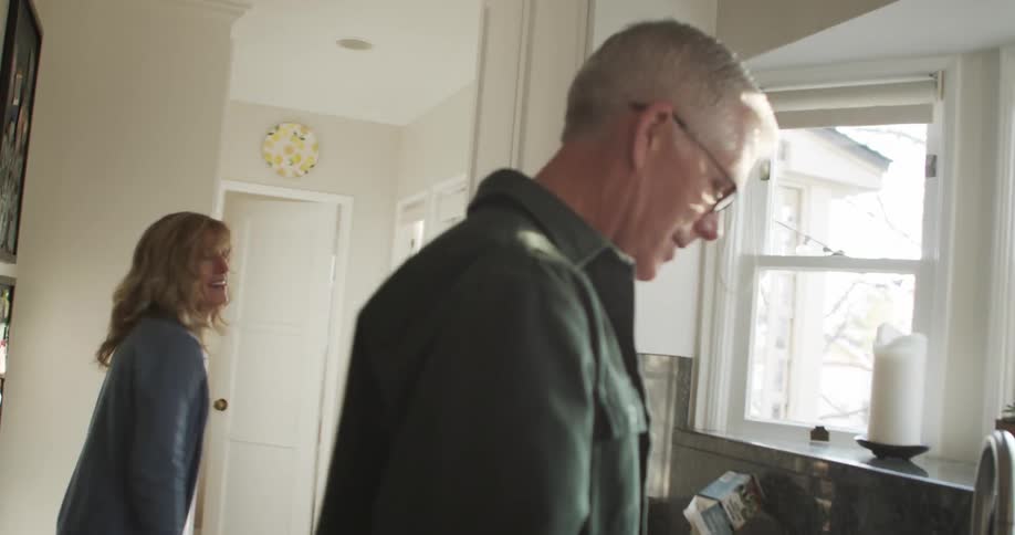 A woman dances around her husband as he washes dishes in their kitchen