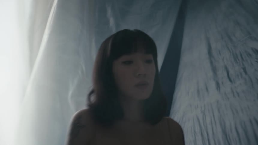 Woman with tattoos stands against blowing translucent fabric staring