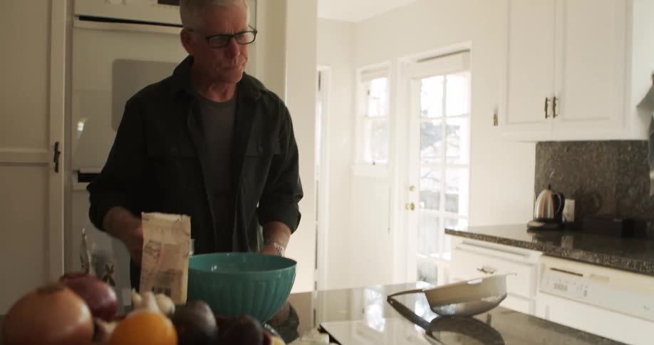 An older male leans over a mixing bowl as he takes out a measuring cup and ingredients