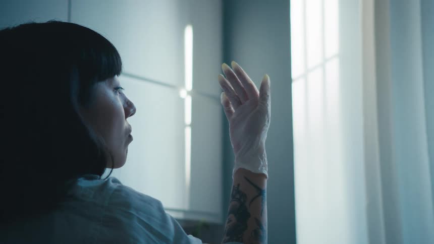 A woman stands in front of a window and stares at her gloved hand