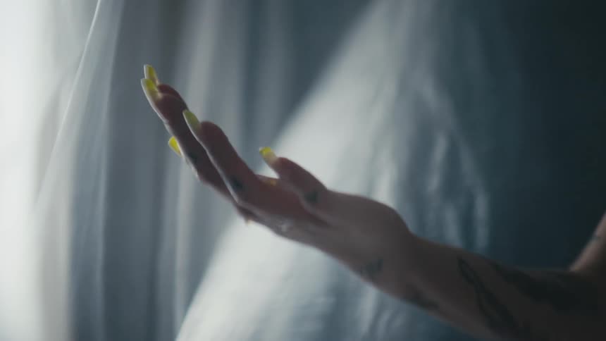 Person with long fingernails painted yellow and tattoo lets water drip through fingers near filmy gray fabric