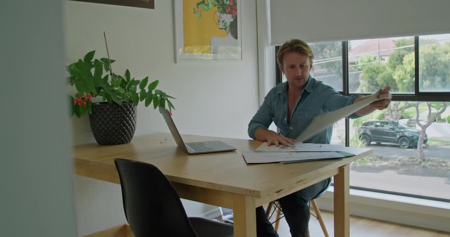 A man goes through blueprints as he sits at his kitchen table with a laptop