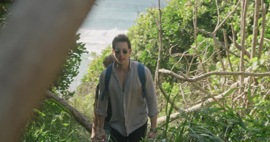 Two men with sunglasses walking up a hill with plants with the ocean in the background