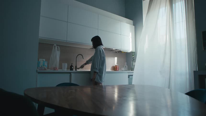 A woman standing in a kitchen next to a sink and faucet with a window in the background