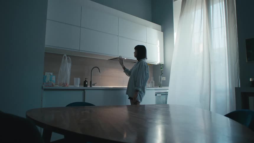 A woman stands in her kitchen and stares blankly at her hand