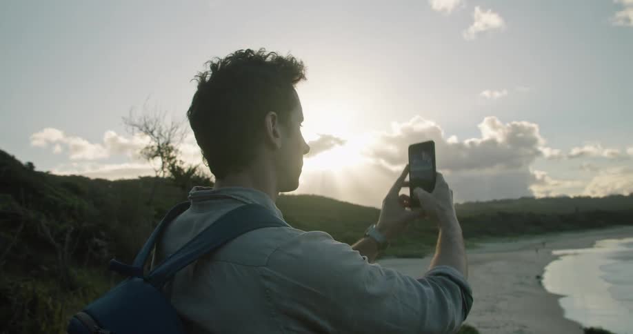 A man standing on a hill uses a smartphone to take a photograph of the beach below before turning and walking away