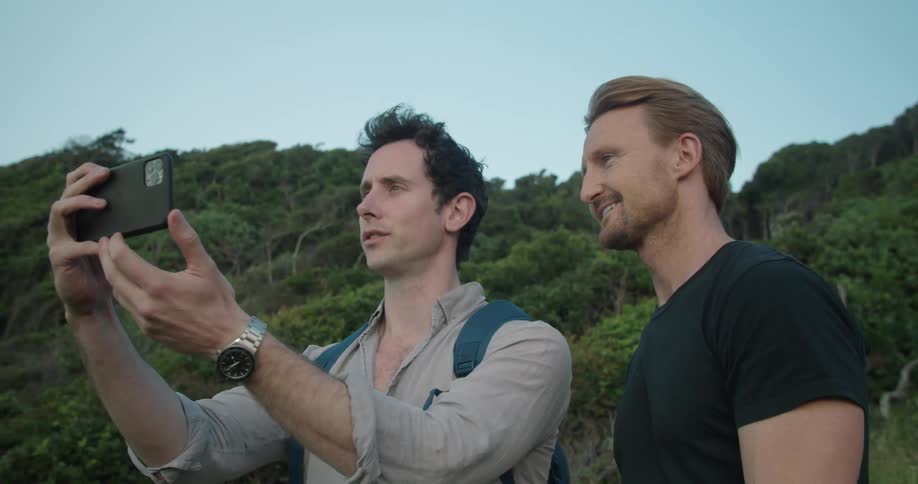 Two men talks near forest covered hill as one takes picture with mobile phone